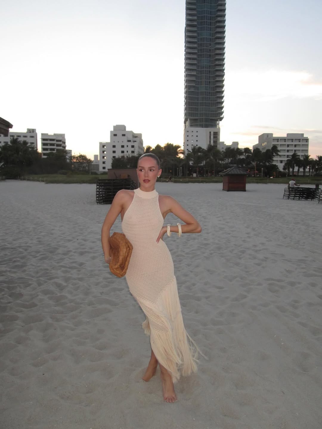 Woman in a long dress standing on a sandy beach with buildings in the background